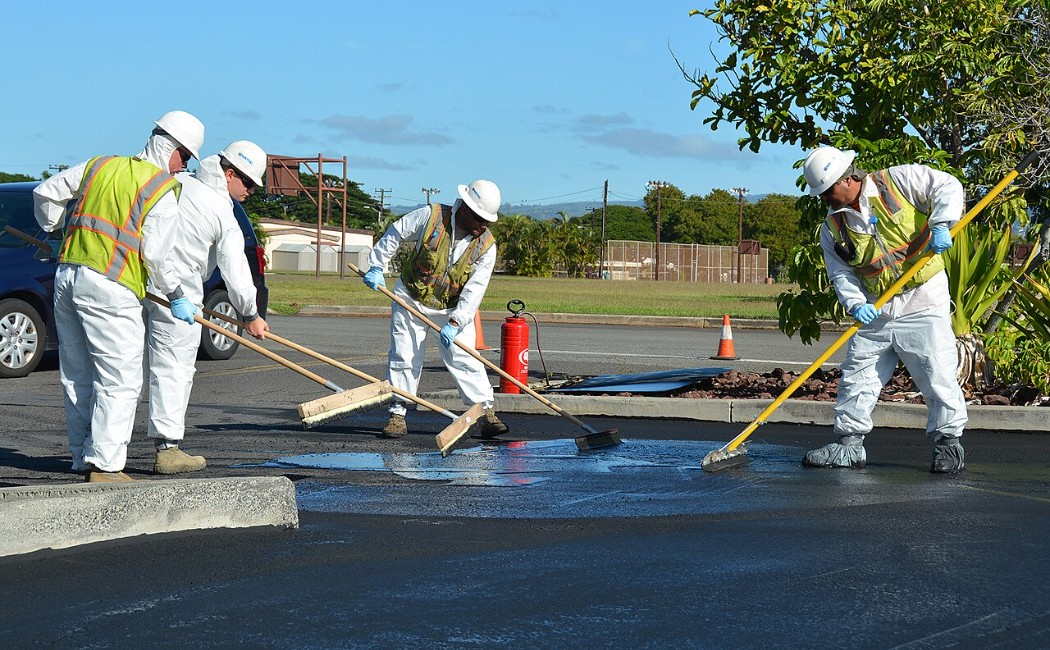 Workers in protective gear applying Sealcoating Services on a parking lot surface using squeegees to spread fresh asphalt sealant.