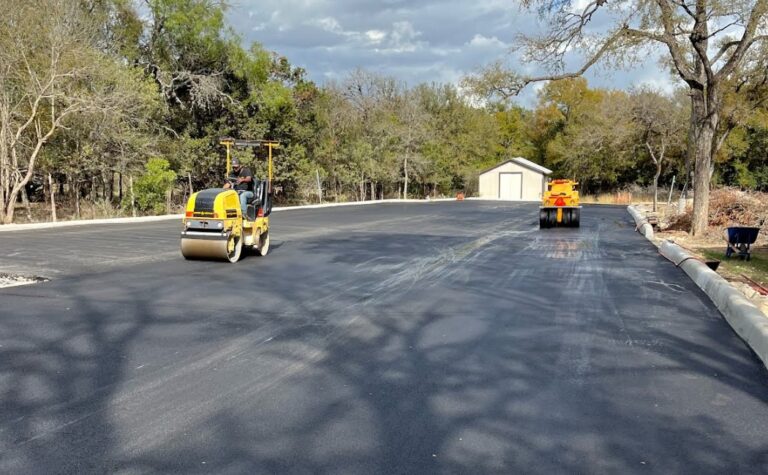 Asphalt Paving in progress with road rollers compacting fresh blacktop on a wide residential driveway surrounded by trees.