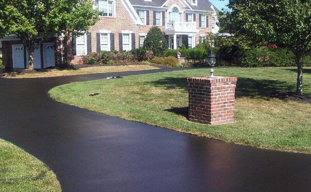 Professional Sealcoating Services for a Smooth, Pristine Driveway Freshly paved driveway with a smooth black finish leading to a large suburban home, showcasing professional Sealcoating Services.