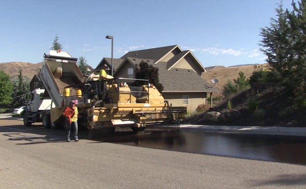 Chip Sealing Services crew applying fresh chip seal coating to a residential neighborhood street with paving equipment and workers directing traffic.