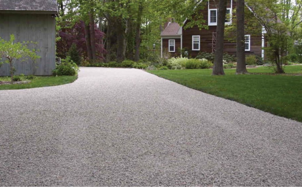 Chip Seal Driveway leading to a residential home, featuring a smooth textured surface bordered by manicured lawns and mature trees.