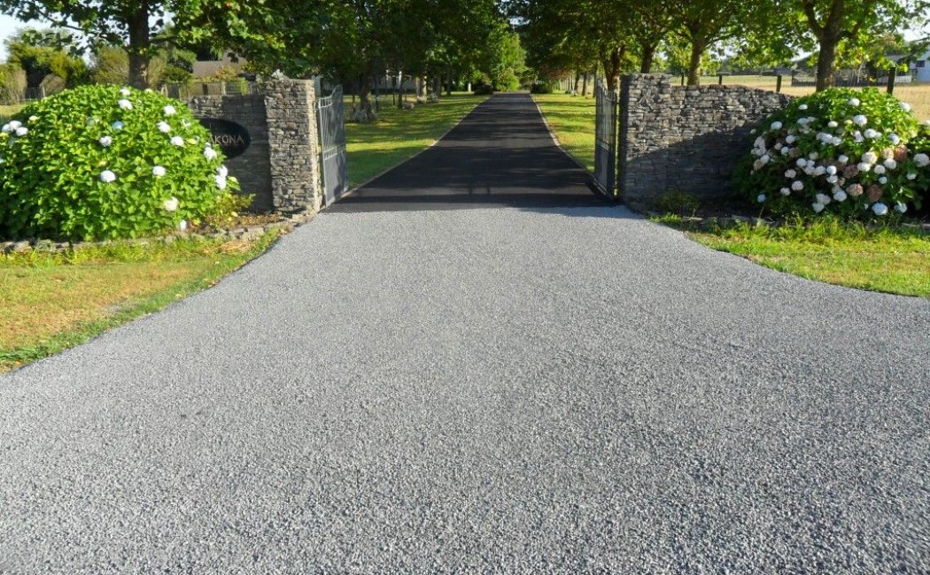Chip Seal Driveway entrance framed by stone walls, flowering shrubs, and a tree-lined lane leading to a scenic countryside property.
