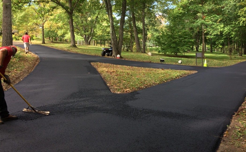 Driveway freshly paved through a wooded residential property, with workers smoothing new asphalt along a branching driveway path.
