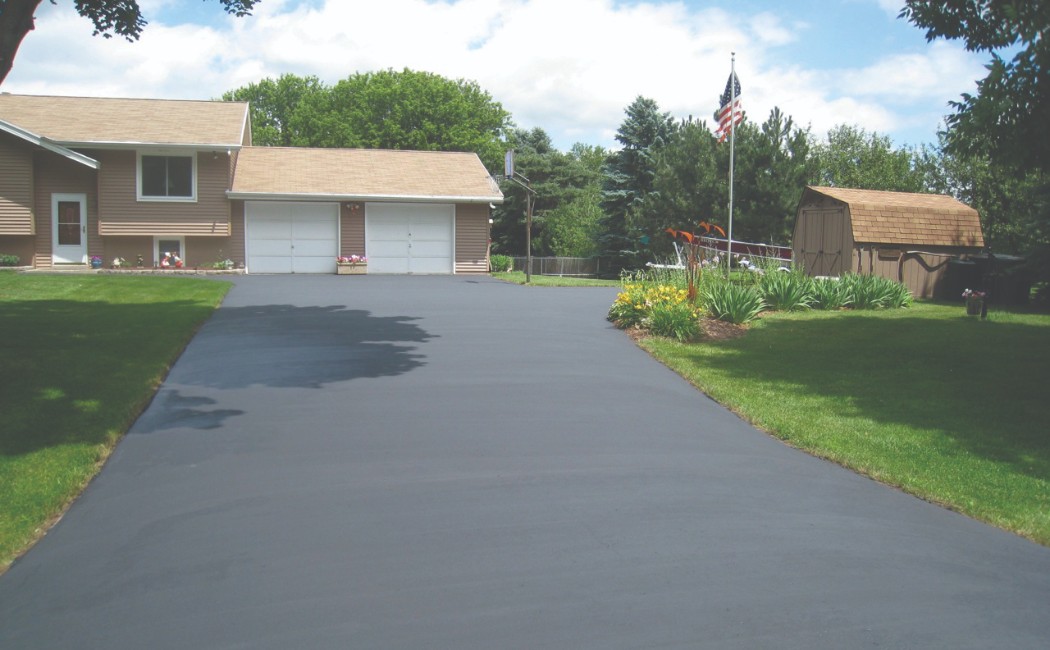 Driveway with a smooth black asphalt surface leading to a suburban home, surrounded by green lawns, landscaping, and mature trees.