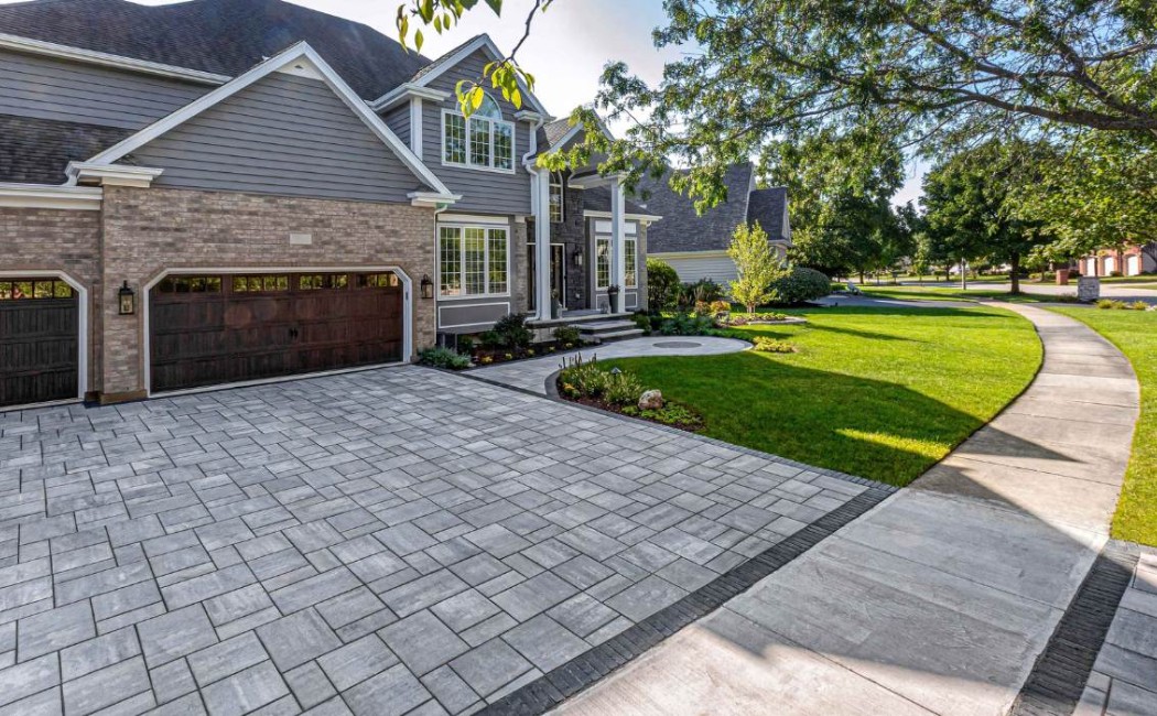 Driveway with elegant interlocking pavers leading to a modern luxury home, surrounded by manicured landscaping and a spacious front yard.