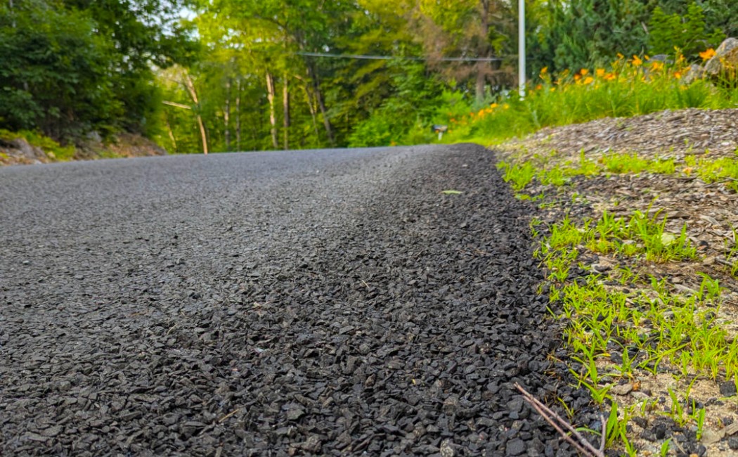 Chip Seal Driveways bordered by fresh grass and trees along a quiet rural road, showcasing a durable and textured paved surface.