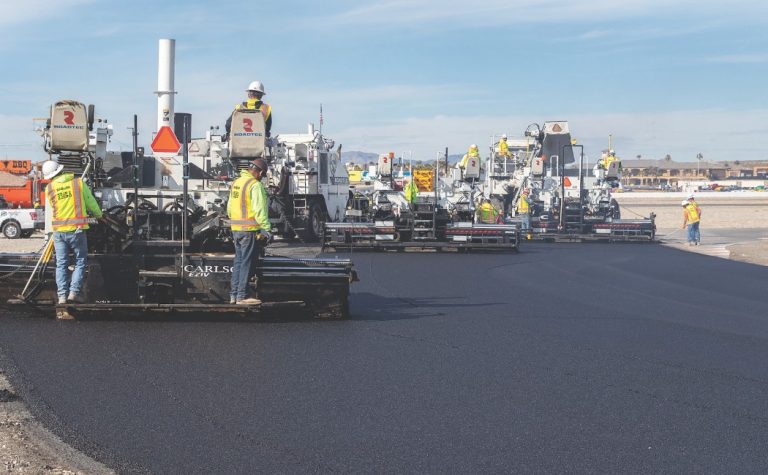 Construction crew performing asphalt paving with multiple paving machines laying fresh black asphalt on a wide roadway construction project.
