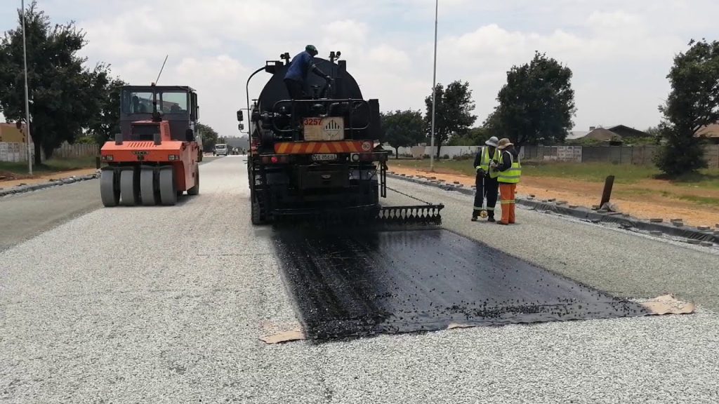 Chip Seal road construction in progress as an asphalt distributor applies bitumen and a road roller compacts the surface, creating a durable, skid-resistant pavement for long-lasting road protection.